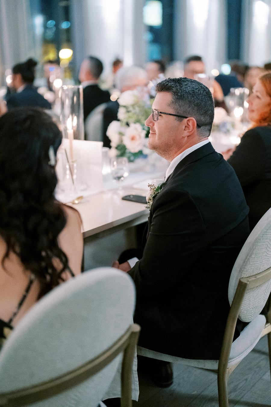 A man in a suit and glasses sitting at a beautifully decorated dinner table during a formal event, surrounded by guests and floral arrangements.