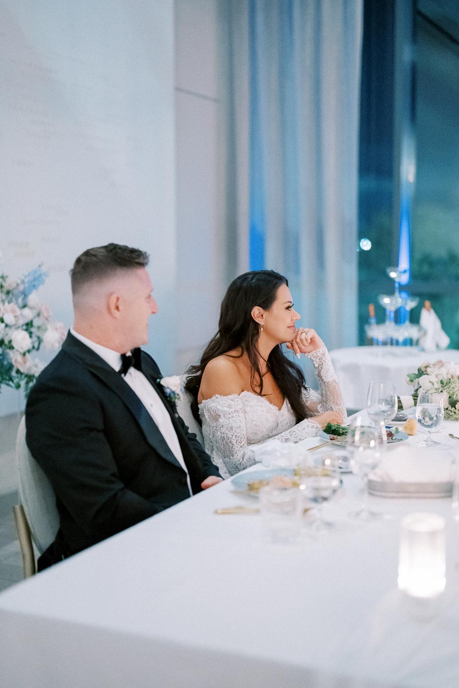 Elegant bride and groom seated at a beautifully decorated wedding reception table, surrounded by ambient lighting and florals, in a romantic celebration atmosphere.