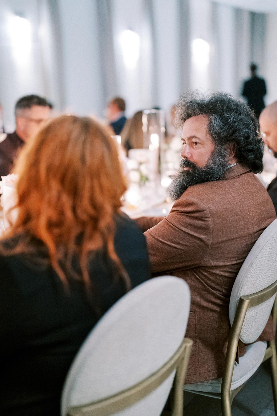 Man with curly hair and beard wearing a brown suit attending a formal dinner event, engaged in conversation amidst elegantly set tables.