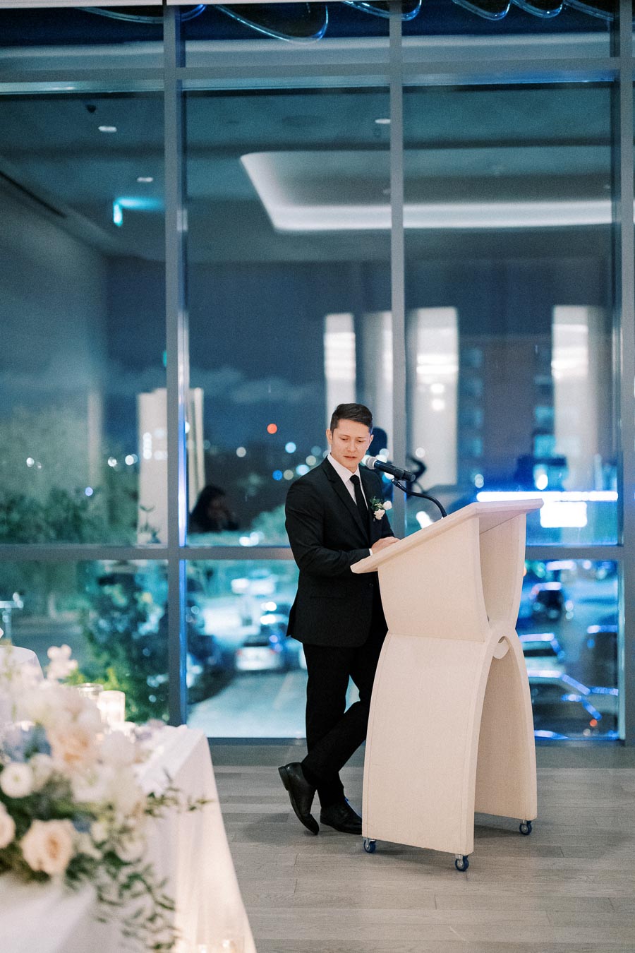A man in a suit giving a speech at a podium indoors, with large windows showing a night cityscape in the background. Flowers and candles decorate the nearby table, suggesting an elegant event setting.