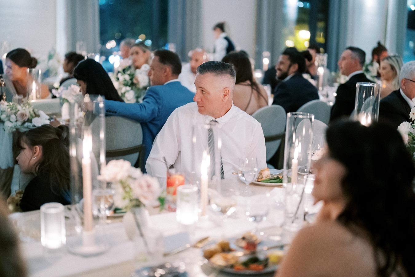 A formal dinner event with elegantly dressed guests seated at tables adorned with floral centerpieces and lit candles, in a softly lit banquet hall.
