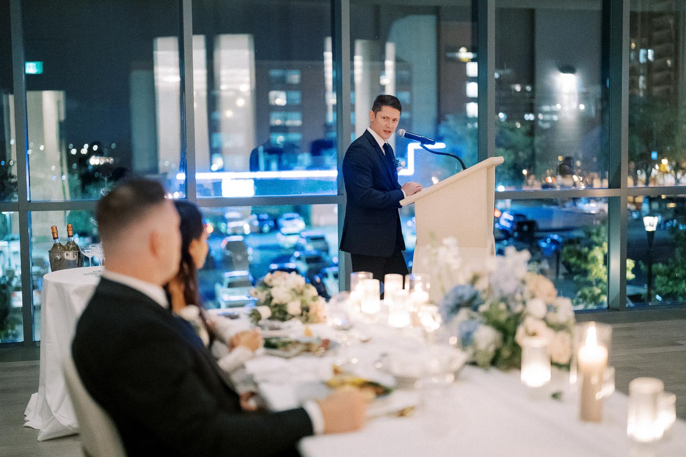 A man in a suit delivers a speech at a wedding reception, standing at a podium with a microphone. The elegantly decorated table with candles and floral arrangements is in the foreground, while blurred guests listen attentively. A large window overlooks a cityscape at night, adding a sophisticated ambiance to the event.
