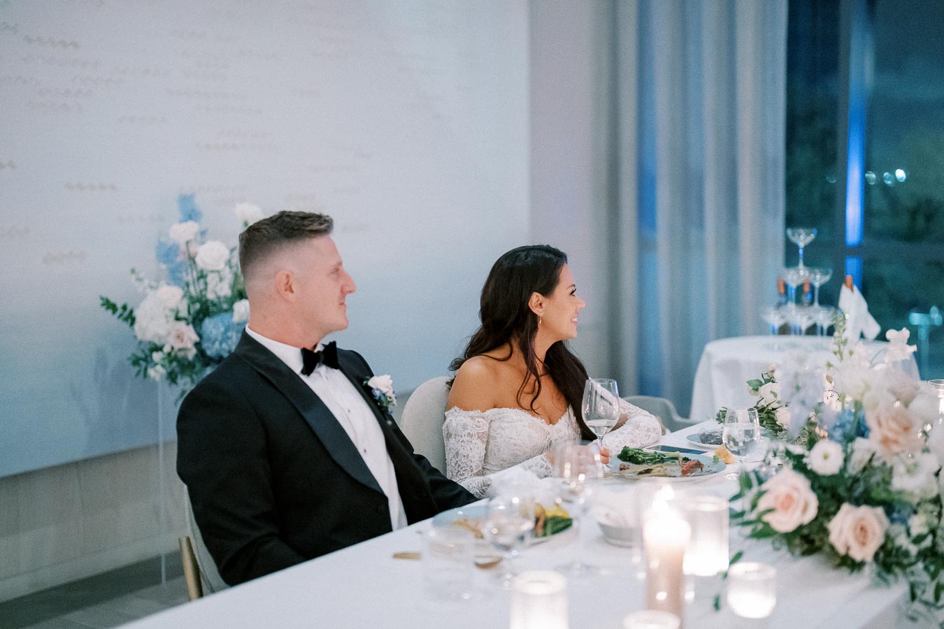 Elegant wedding reception with a bride in a lace off-the-shoulder gown and groom in a black tuxedo seated at a beautifully decorated table with floral arrangements and candles.