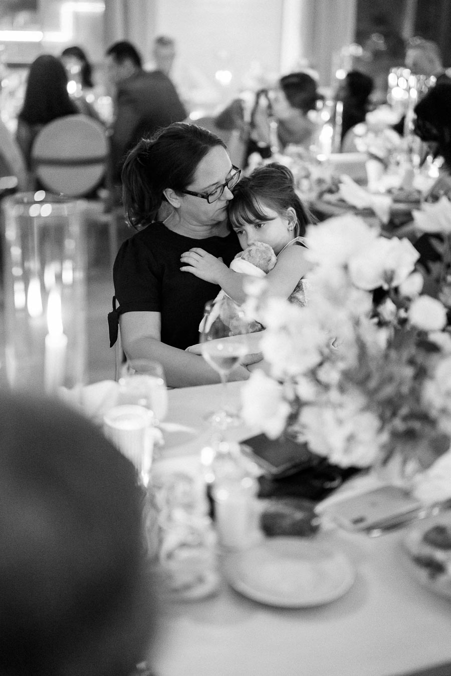 Black and white image of a woman embracing a young girl in a warmly lit, elegant dining setting with blurred guests and floral arrangements in the background.