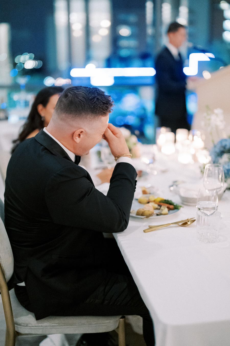 A man in a formal suit sits at a dining table, covering his face with his hand in emotion during a wedding speech, with a blurred background of a speaker and candlelit decor.