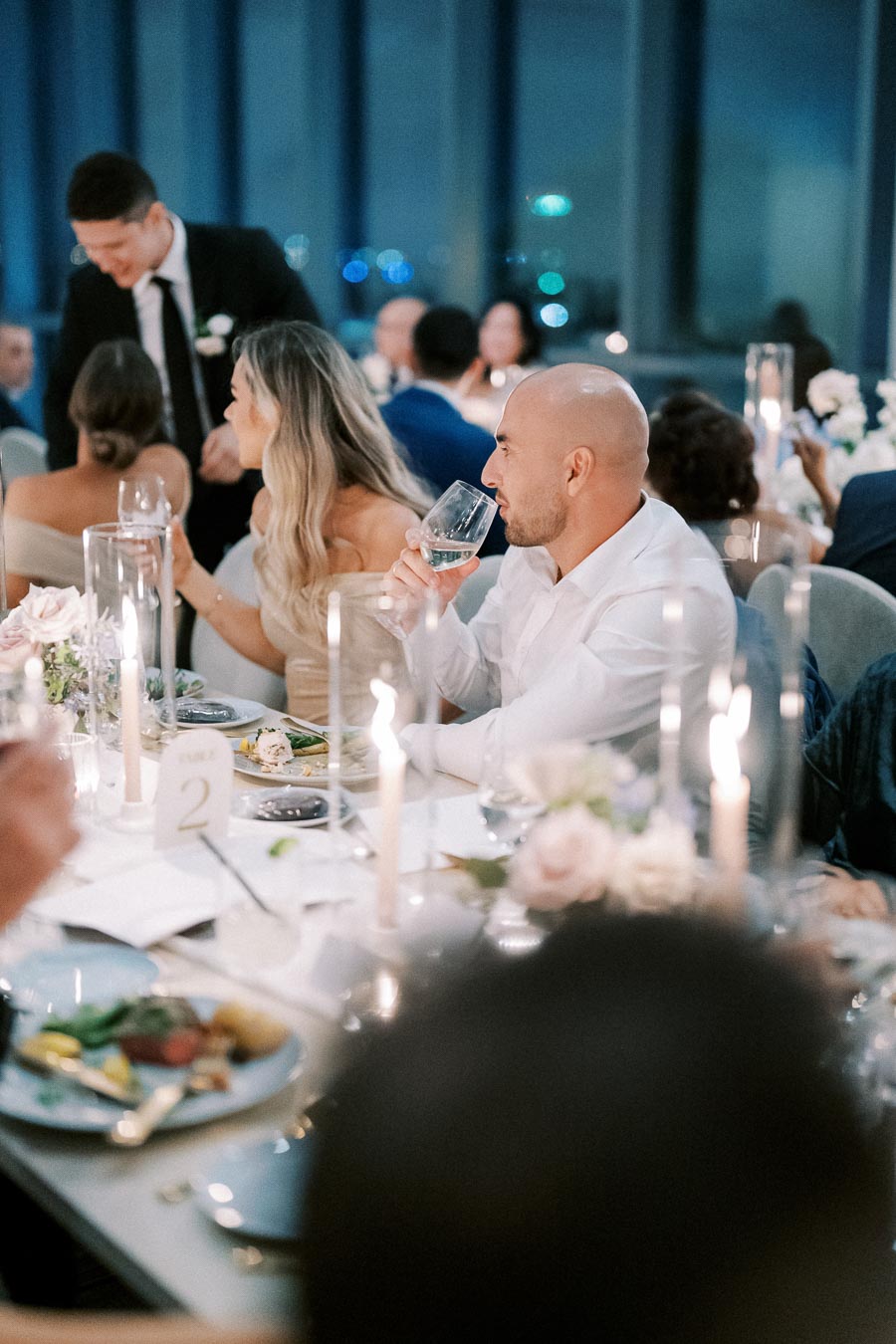 A group of elegantly dressed guests enjoying a candlelit wedding reception dinner, with floral centerpieces and wine glasses on the table.