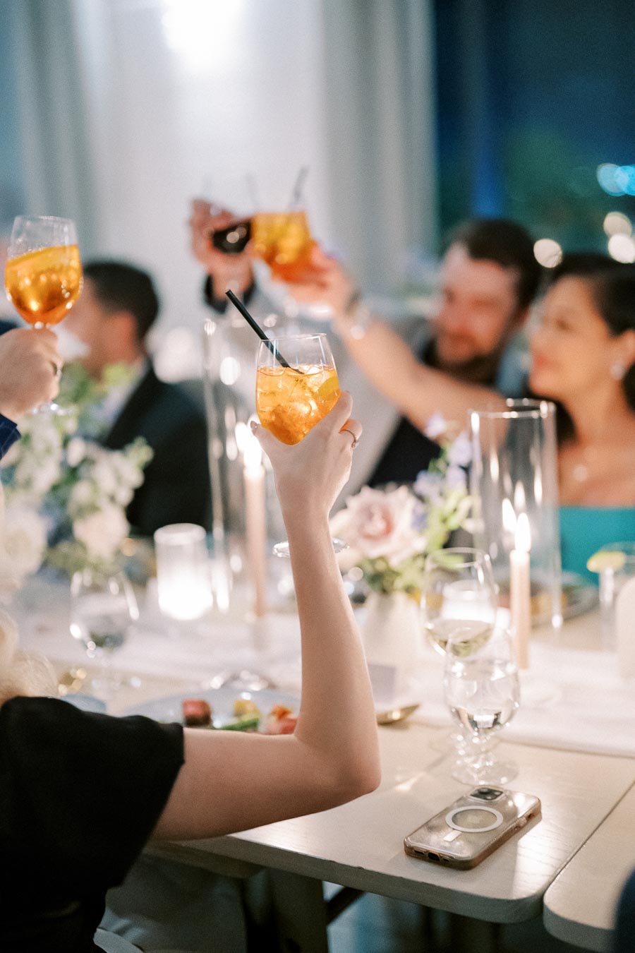 Guests raising glasses of orange cocktails in a celebratory toast at an elegant dinner party, with a table set with candles and floral arrangements.