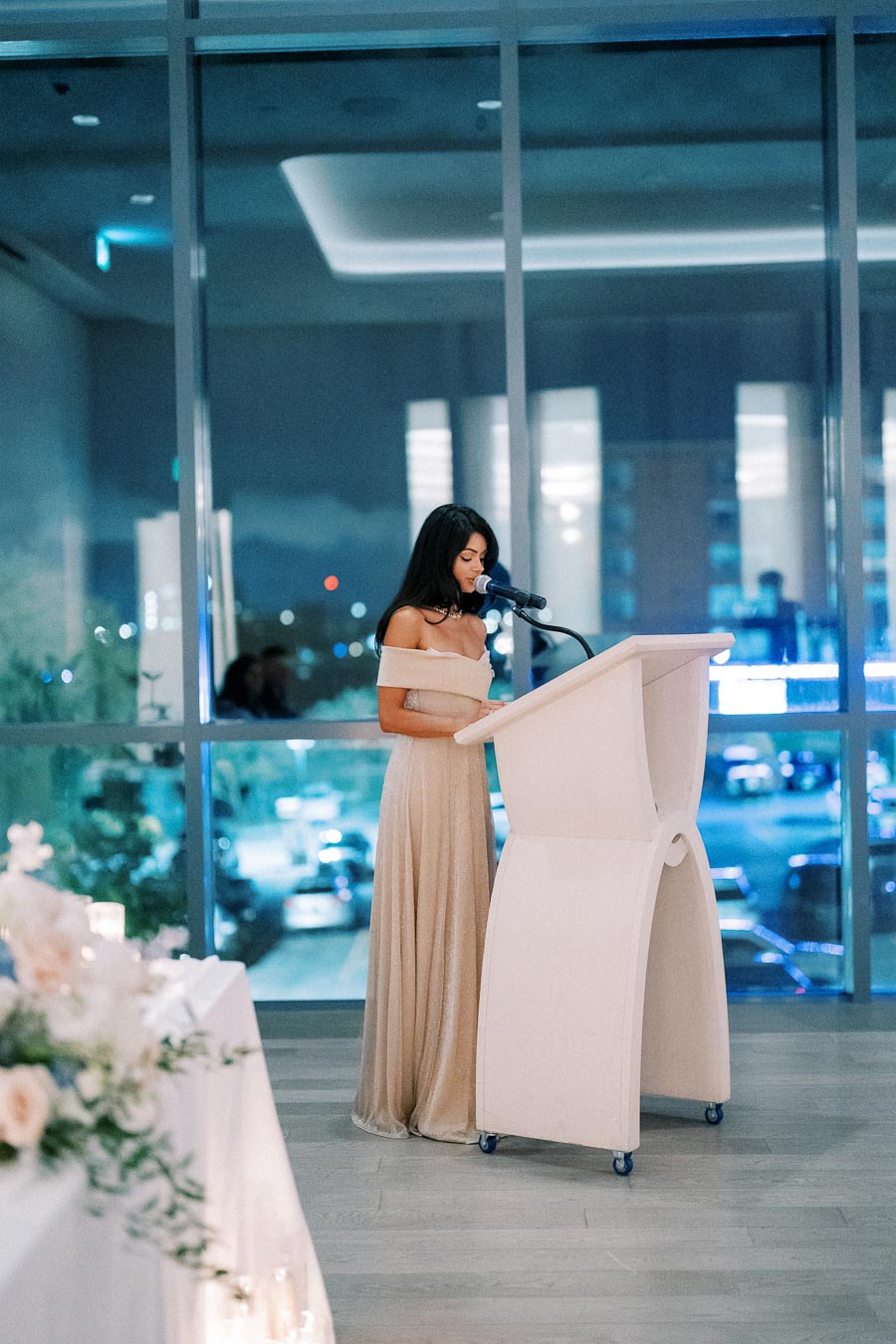 Elegant woman in an evening gown giving a speech at a podium in a modern venue with large windows showcasing city lights at night.