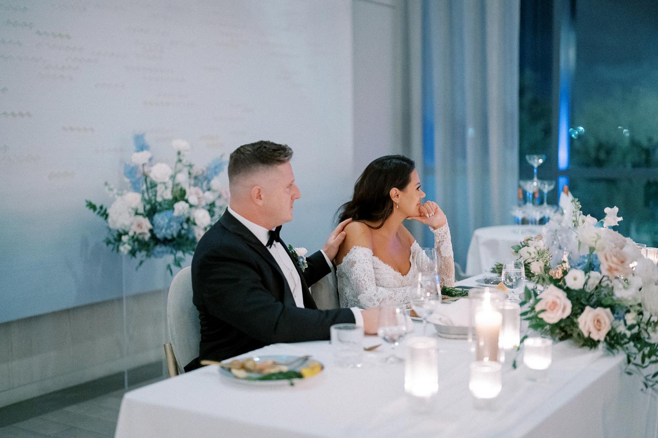 Wedding reception scene with a bride in an elegant lace gown and a groom in a black tuxedo seated at a beautifully decorated table, adorned with white and blush floral arrangements and candlelight.