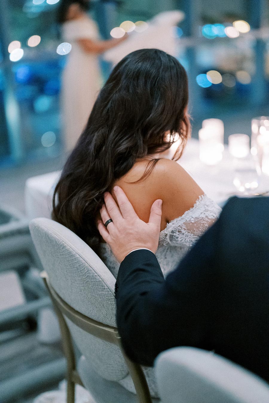 A romantic moment is captured as a man gently places a hand on a woman's shoulder at an elegant dinner event. They are seated at a table adorned with glowing candles, creating a warm and intimate atmosphere.