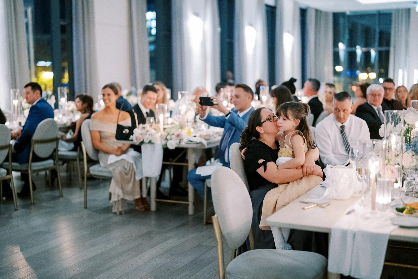 A warm and elegant wedding reception in a modern venue, featuring guests seated at beautifully adorned tables with white floral centerpieces and candles. A mother shares a joyful moment, embracing her young daughter, while other attendees smile and enjoy the ambiance.