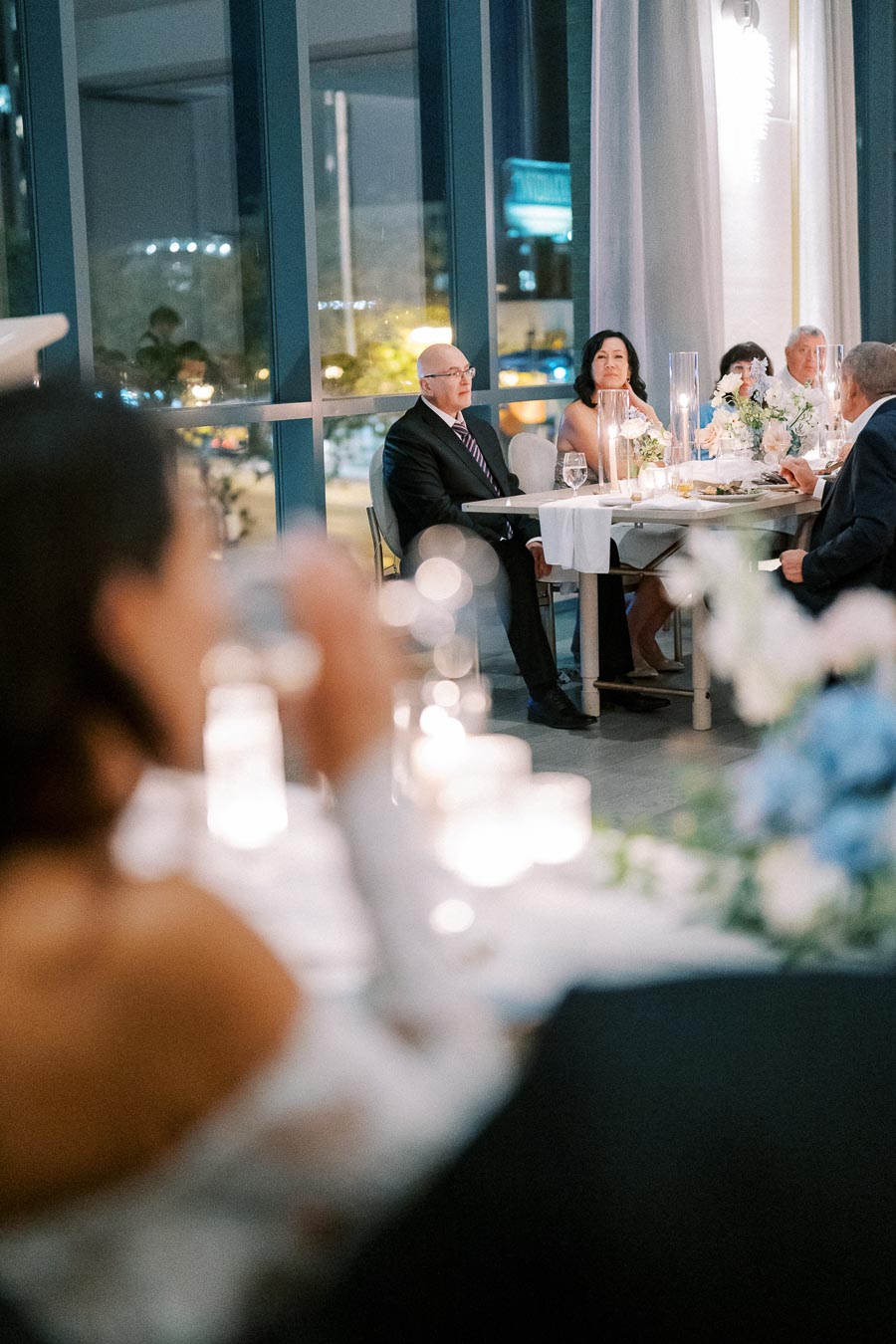 Elegant indoor wedding reception with guests seated at a beautifully decorated table, featuring white floral arrangements and candlelight, set against large windows with a nighttime city view.