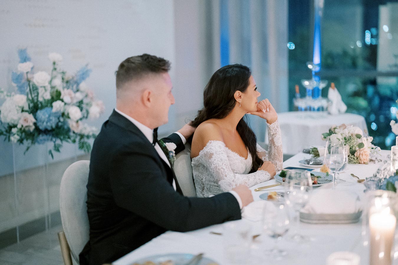 Alt text: Elegant couple seated at a wedding reception, bride in off-shoulder lace gown and groom in black tuxedo, surrounded by floral arrangements and candlelit table setting.