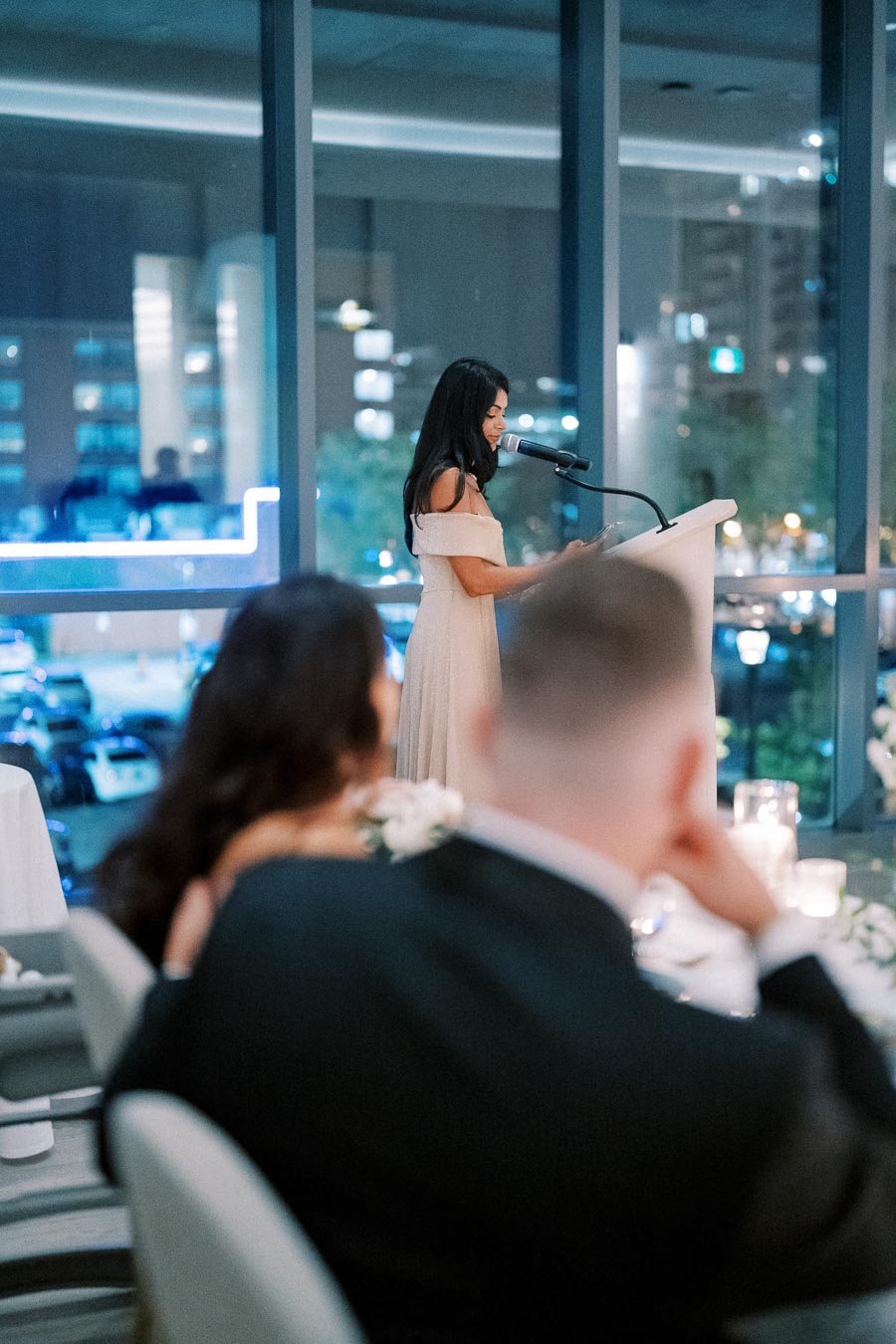 A woman in a formal dress delivers a speech at a wedding reception. She stands at a podium with a microphone, while guests seated nearby listen attentively. The event is held in a modern venue with large windows showcasing a city view at night.