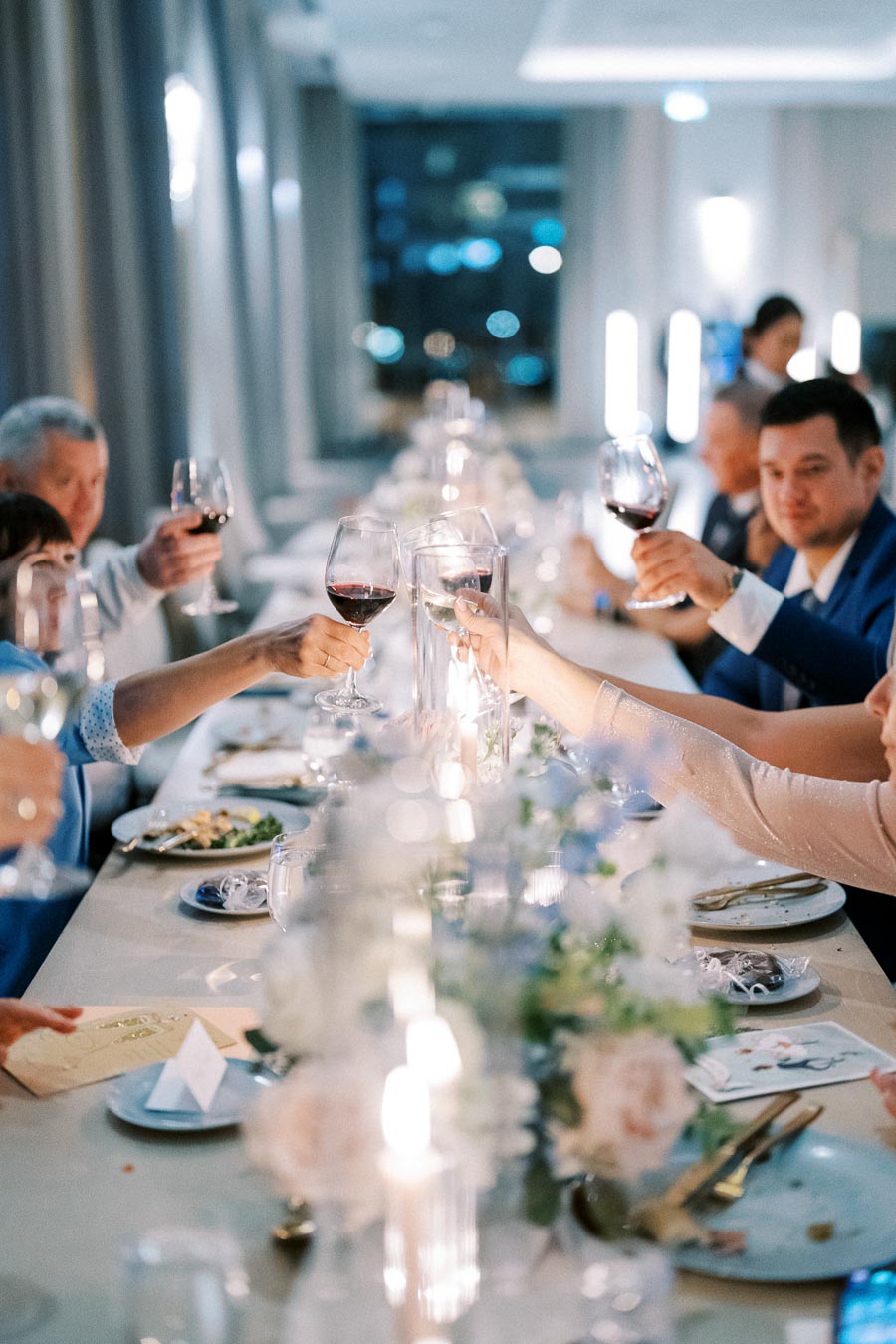 Elegant dinner gathering with guests toasting wine glasses at a beautifully decorated table, featuring a soft focus on plates and floral arrangements, creating a festive atmosphere.