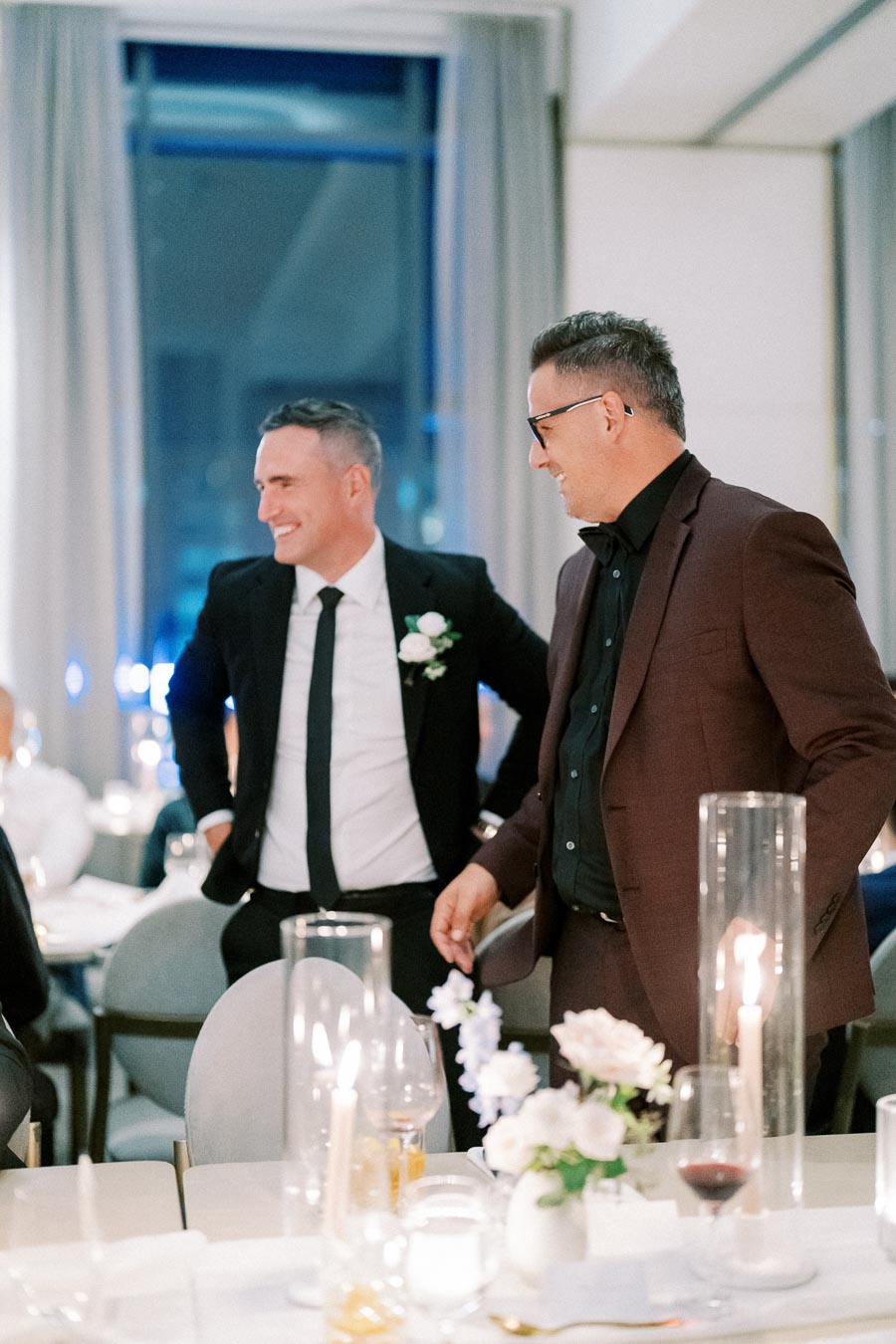 Two men in suits smiling and talking at an elegant event, surrounded by candlelit tables with floral arrangements.