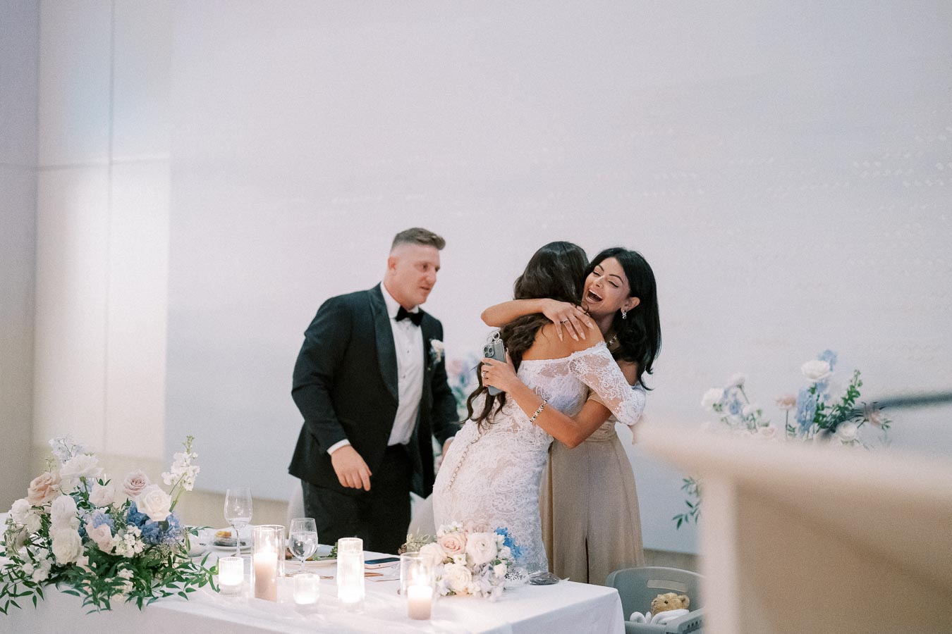 Three people celebrating at a wedding reception; two women embrace joyfully near a decorated table with floral arrangements and candles, while a man in a tuxedo stands nearby.