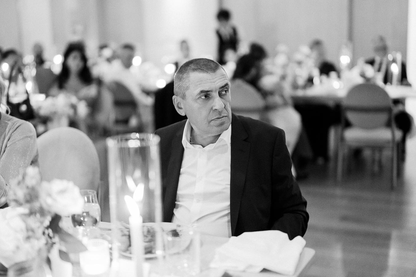 A man in formal attire sitting at an elegantly set dinner table, surrounded by guests in a warmly lit banquet hall.
