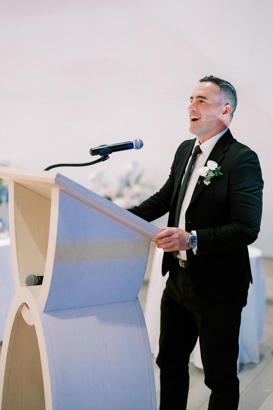 A man in a suit smiling confidently while speaking at a podium with a microphone during a formal event.