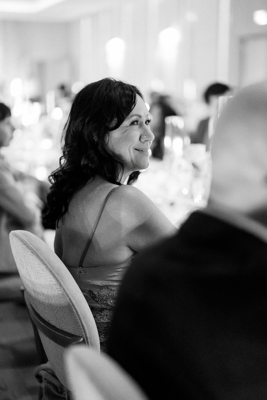 Black and white photo of a smiling woman with dark hair sitting at an elegant event, wearing a dress with delicate detailing, blurred background.