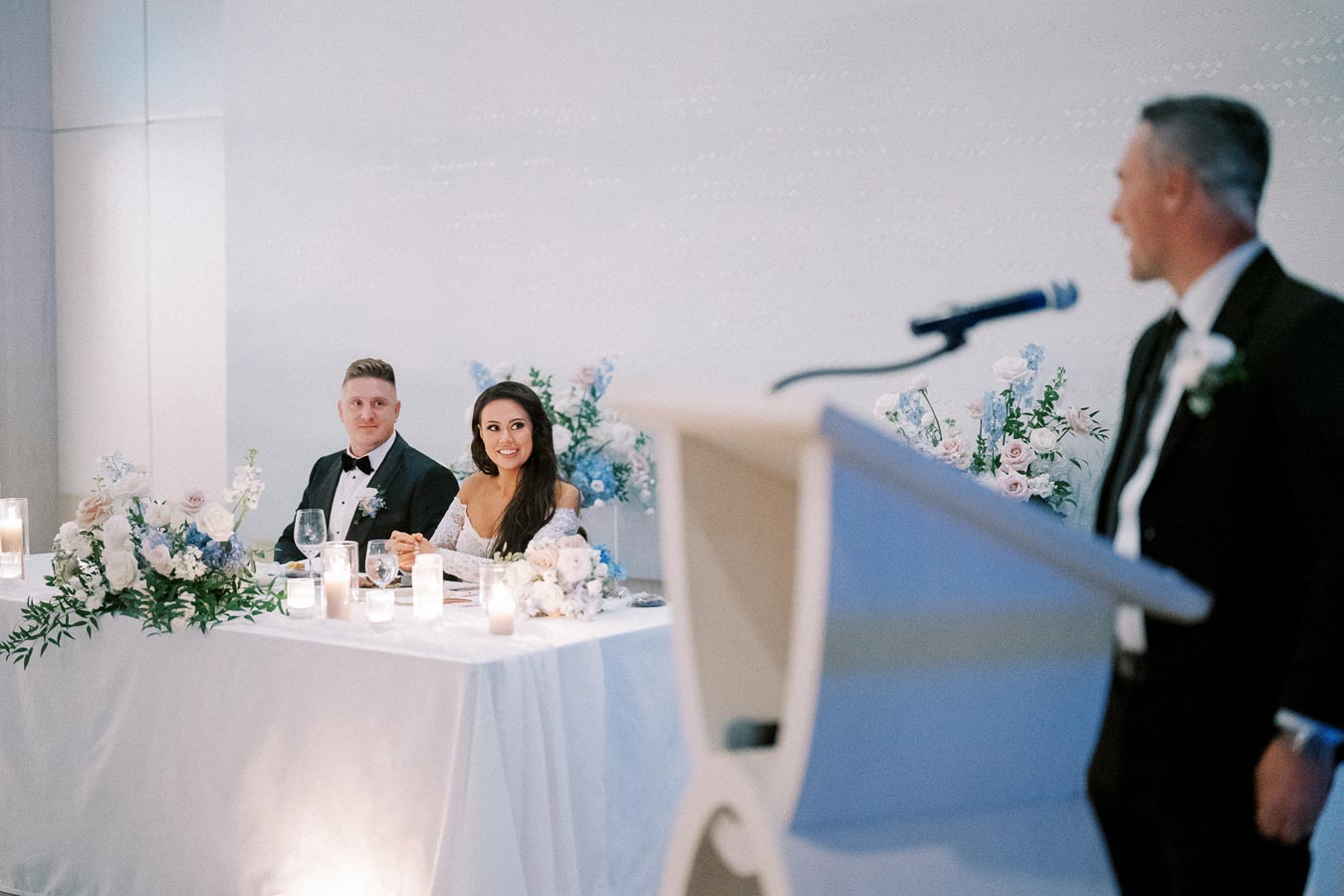 Wedding reception scene with a bride and groom sitting at a decorated table, listening to a speech. The table is adorned with candles and floral arrangements. A speaker is blurred in the foreground, standing at a podium.