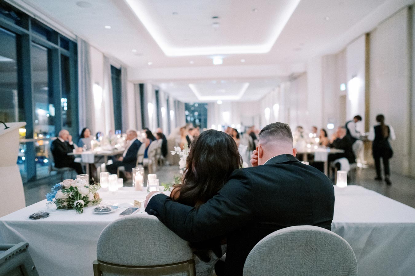 A couple seated together at a beautifully decorated wedding reception, surrounded by guests in an elegant, modern venue with soft lighting and floral arrangements.