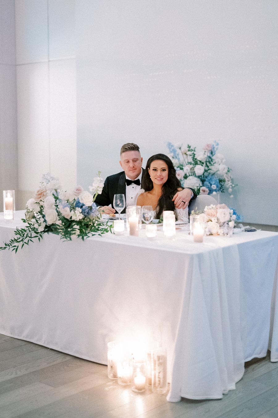 Elegant wedding reception table with a couple seated, adorned with white and blue floral arrangements, candles, and a white tablecloth in a modern venue.