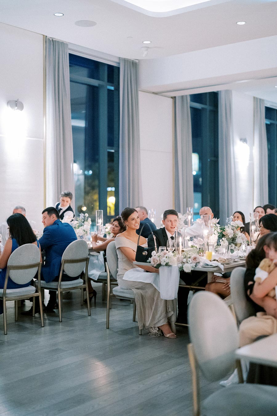 Elegant indoor wedding reception with guests seated at tables, adorned with floral centerpieces and candles, in a modern venue with large windows and soft lighting.