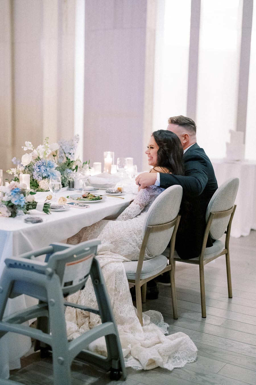 A newlywed couple sits closely together at an elegantly decorated wedding reception table, adorned with floral arrangements and candles, creating a romantic atmosphere.