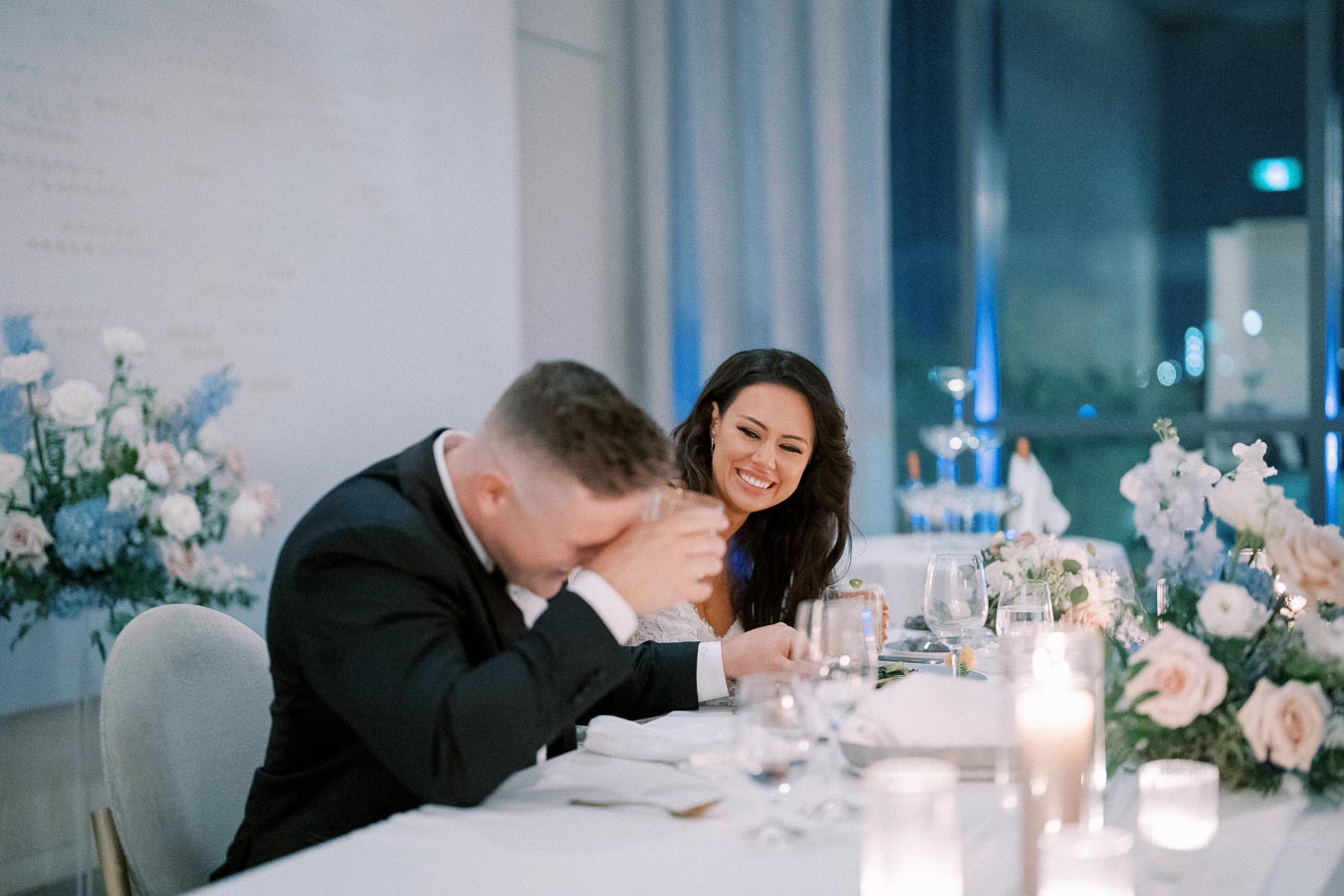 A couple laughing and enjoying a wedding reception at a beautifully decorated table with flowers and candles.