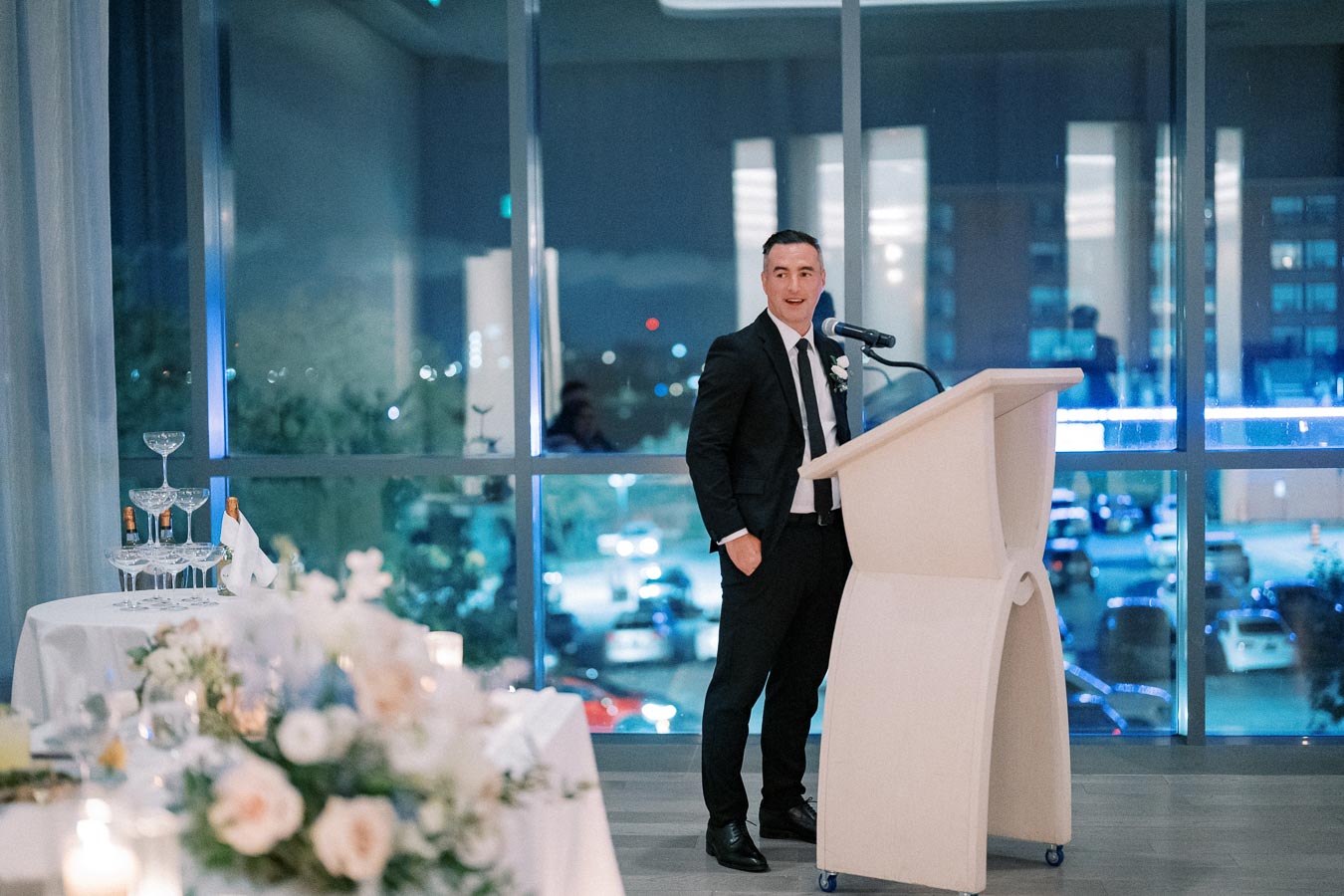 A man in a suit stands at a podium delivering a speech at a formal event, with a champagne tower and floral decorations visible on a nearby table.
