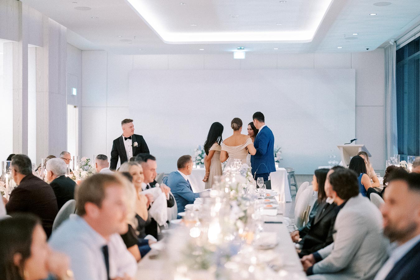 A beautifully decorated wedding reception hall with elegantly dressed guests seated at tables adorned with candles and floral centerpieces, creating a warm and inviting atmosphere.