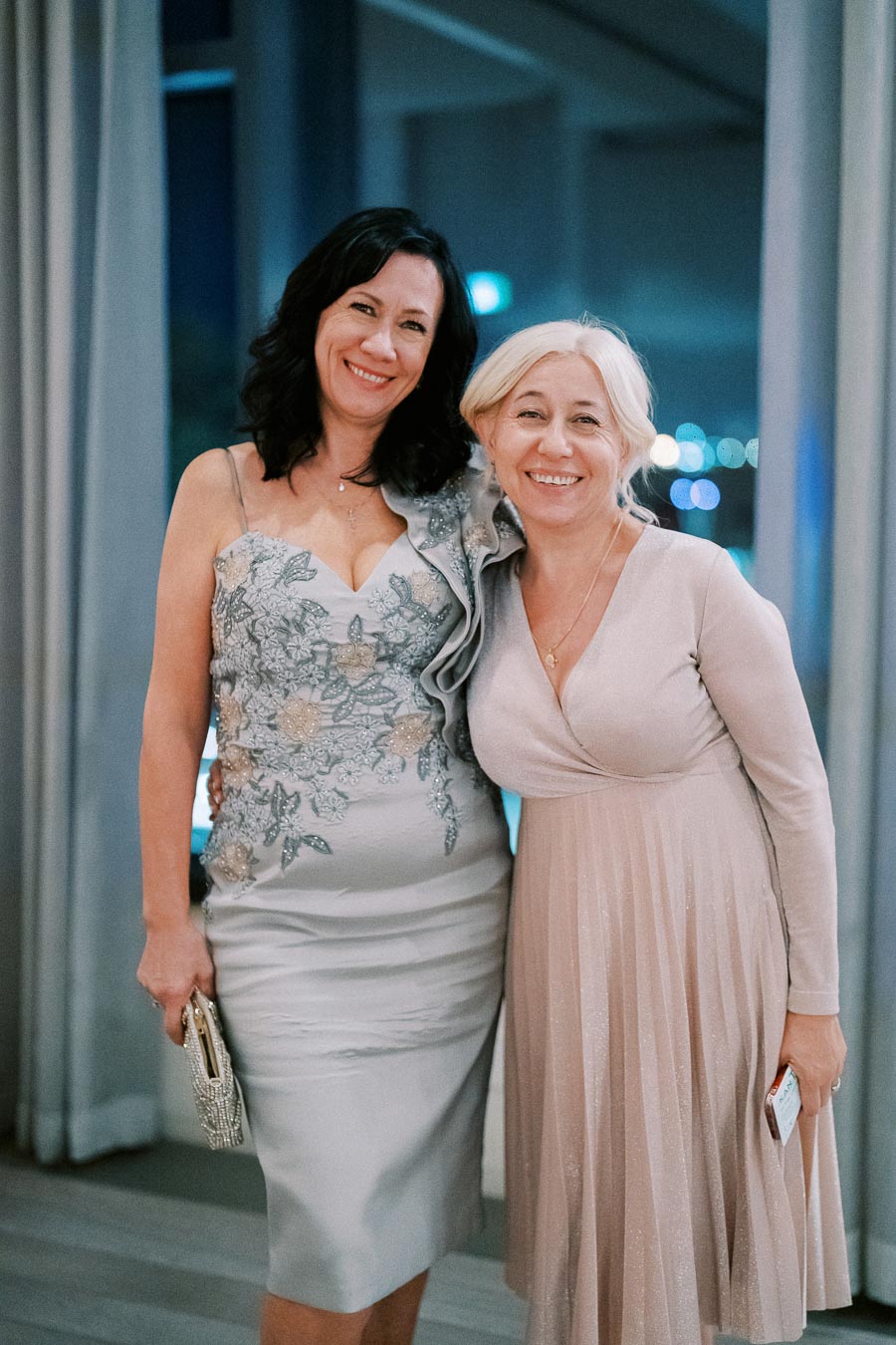 Two women in elegant dresses smiling and posing together indoors at an event.