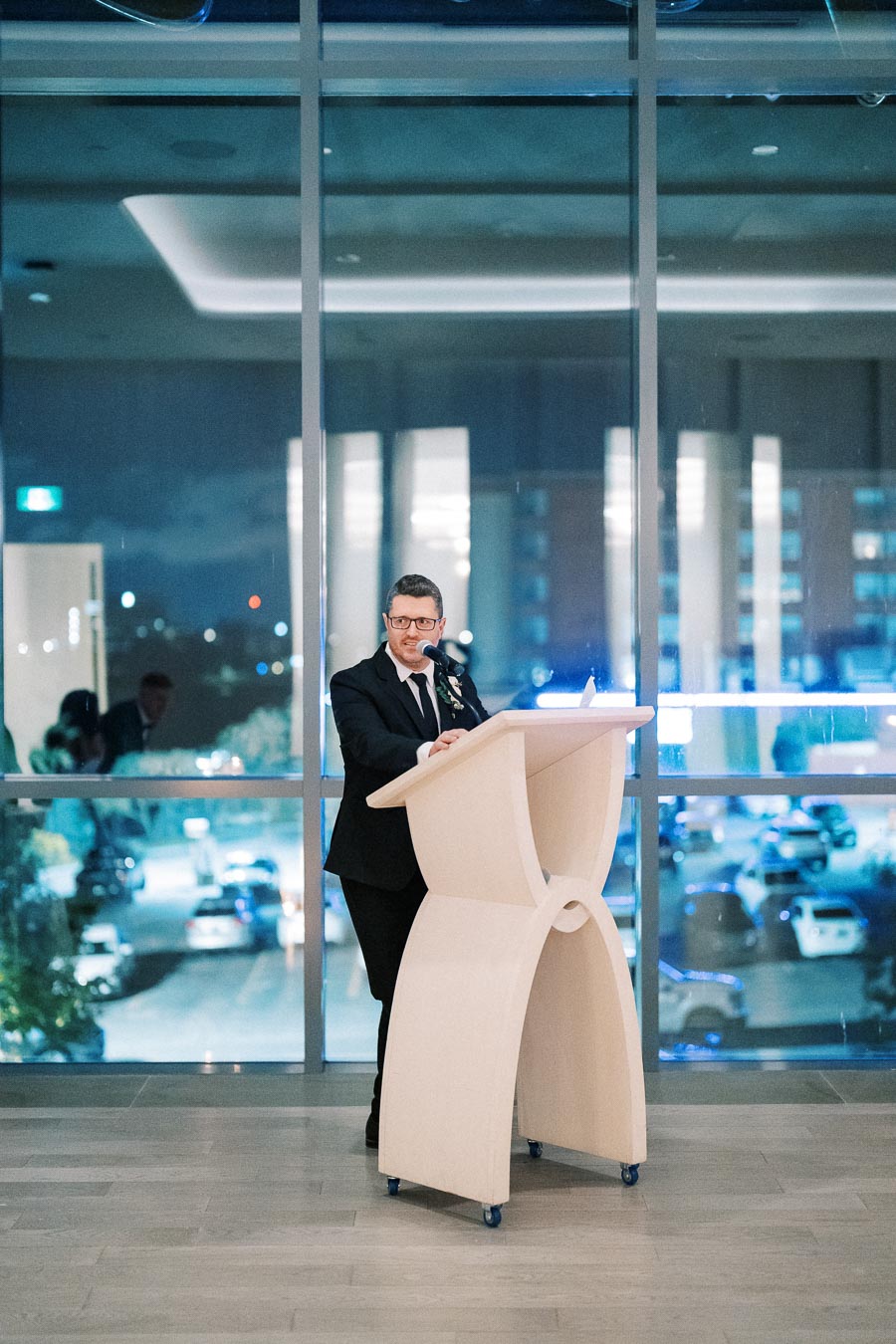 Man in a suit delivering a speech at a podium indoors, with a nighttime city view through the large glass window behind him.
