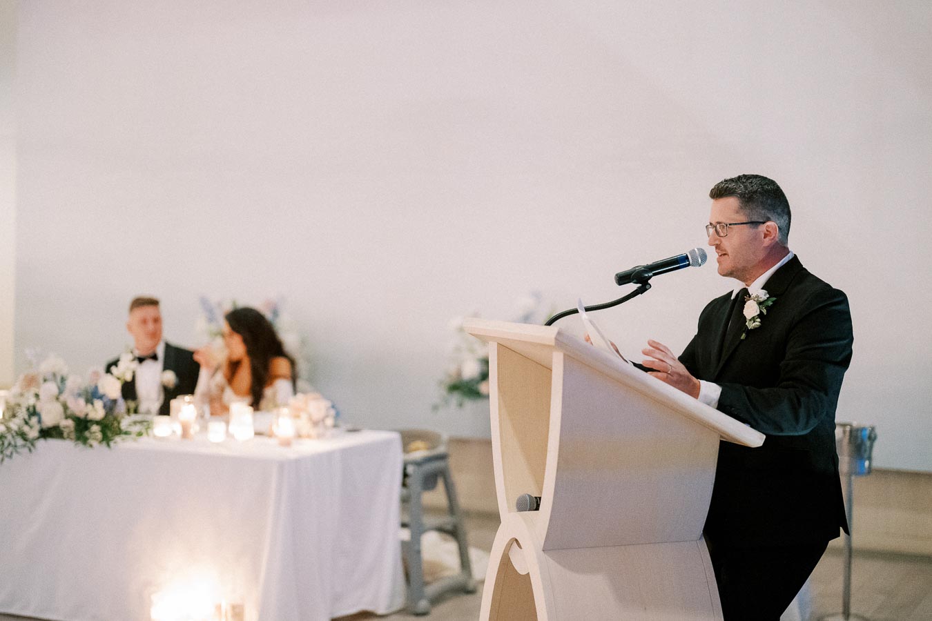 A man in a suit gives a speech at a wedding reception, speaking into a microphone at a podium. In the background, a bride and groom sit at a table adorned with flowers and candles.