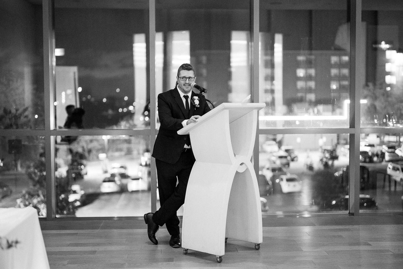 A person giving a speech at a podium in a formal setting, with a cityscape visible through large windows in the background.