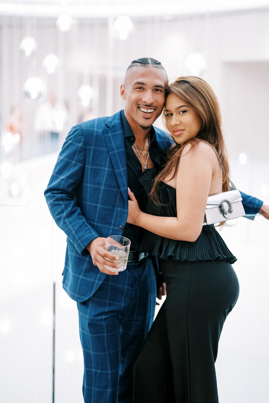 A stylish couple smiling and posing for a photo at an elegant event; the man in a blue plaid suit holding a drink, and the woman in a black dress with a white purse.