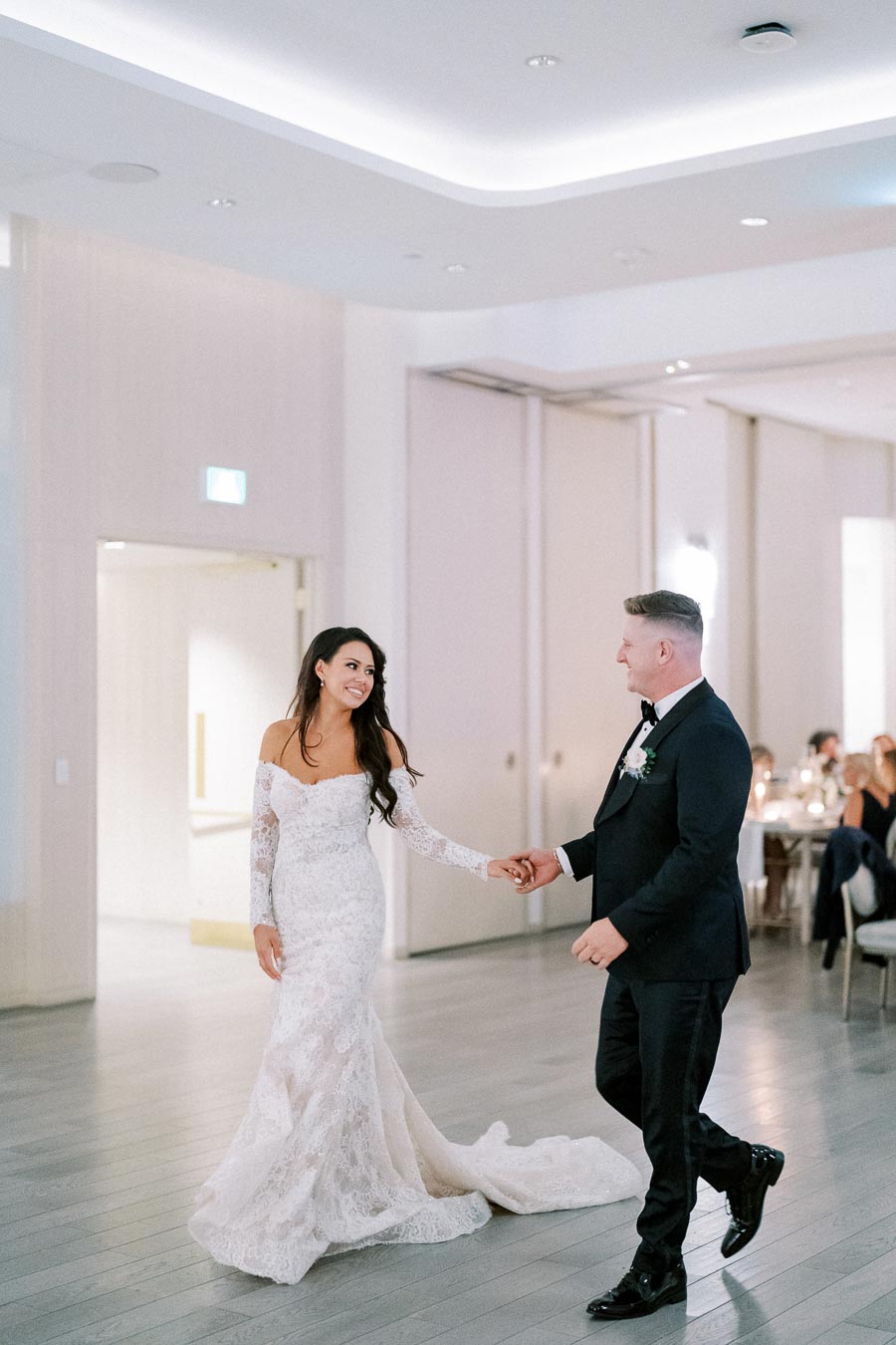 Wedding couple dancing in elegant reception hall, bride in lace gown smiling at groom in black suit and bow tie.