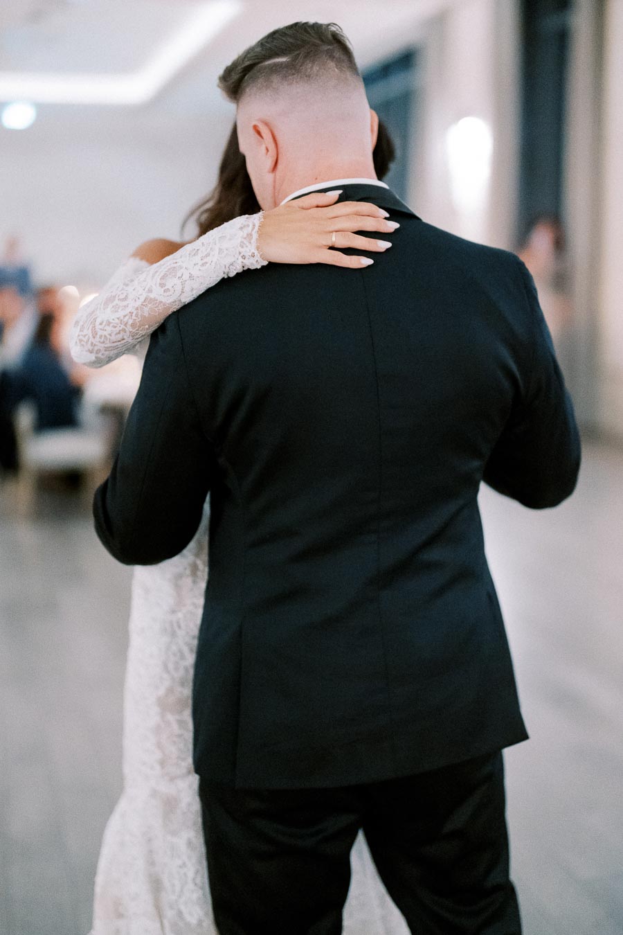 A couple sharing a romantic dance at their wedding, with the bride in a lace dress embracing the groom in a suit, captured in a softly lit venue.