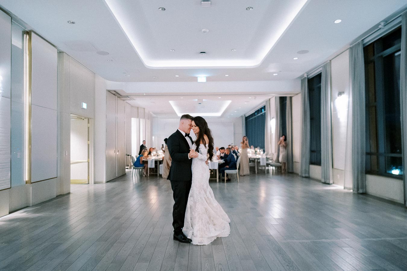 Elegant wedding reception with bride and groom sharing first dance in a beautifully lit ballroom, surrounded by guests seated at dining tables.