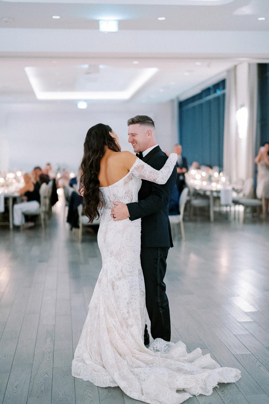 A bride and groom sharing their first dance in a beautifully decorated wedding hall, with the bride in a lace gown and the groom in a black suit, surrounded by seated guests and softly lit candles.