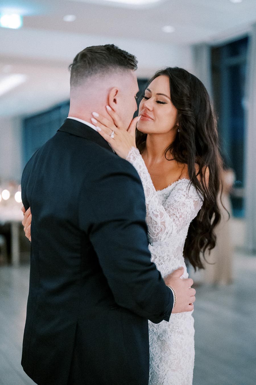 A bride and groom share a romantic first dance at their wedding reception, with the bride in a lace gown holding her groom's face lovingly.