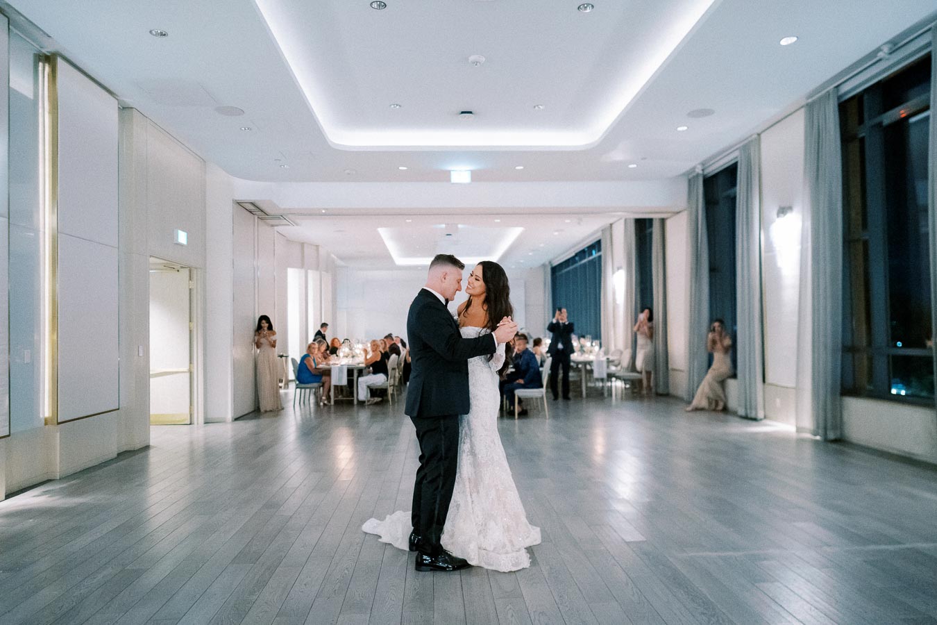 Elegant wedding reception with newlywed couple sharing first dance in a beautifully decorated modern venue, surrounded by guests seated at round tables.