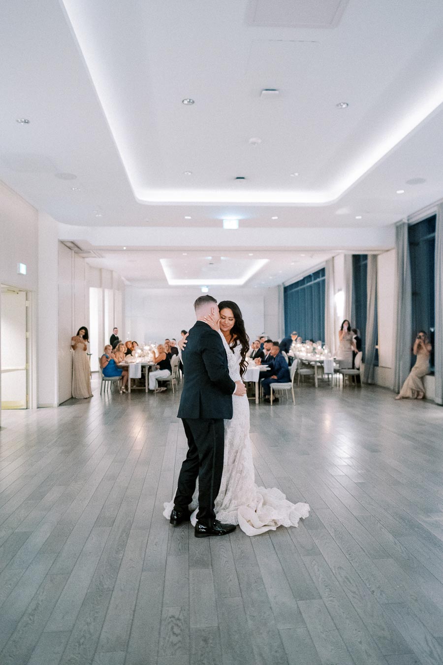 A bride and groom share their first dance in an elegantly decorated wedding reception hall with guests seated at tables.