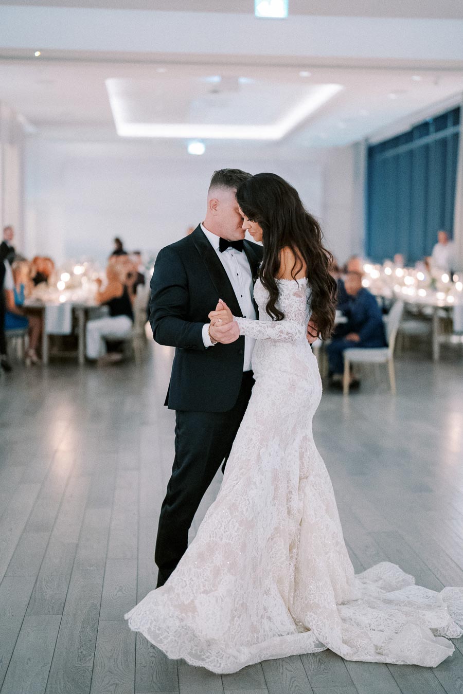 Elegant couple dancing at a wedding reception, with the bride in a stunning lace gown and the groom in a classic black tuxedo, surrounded by softly lit tables creating a romantic ambiance.
