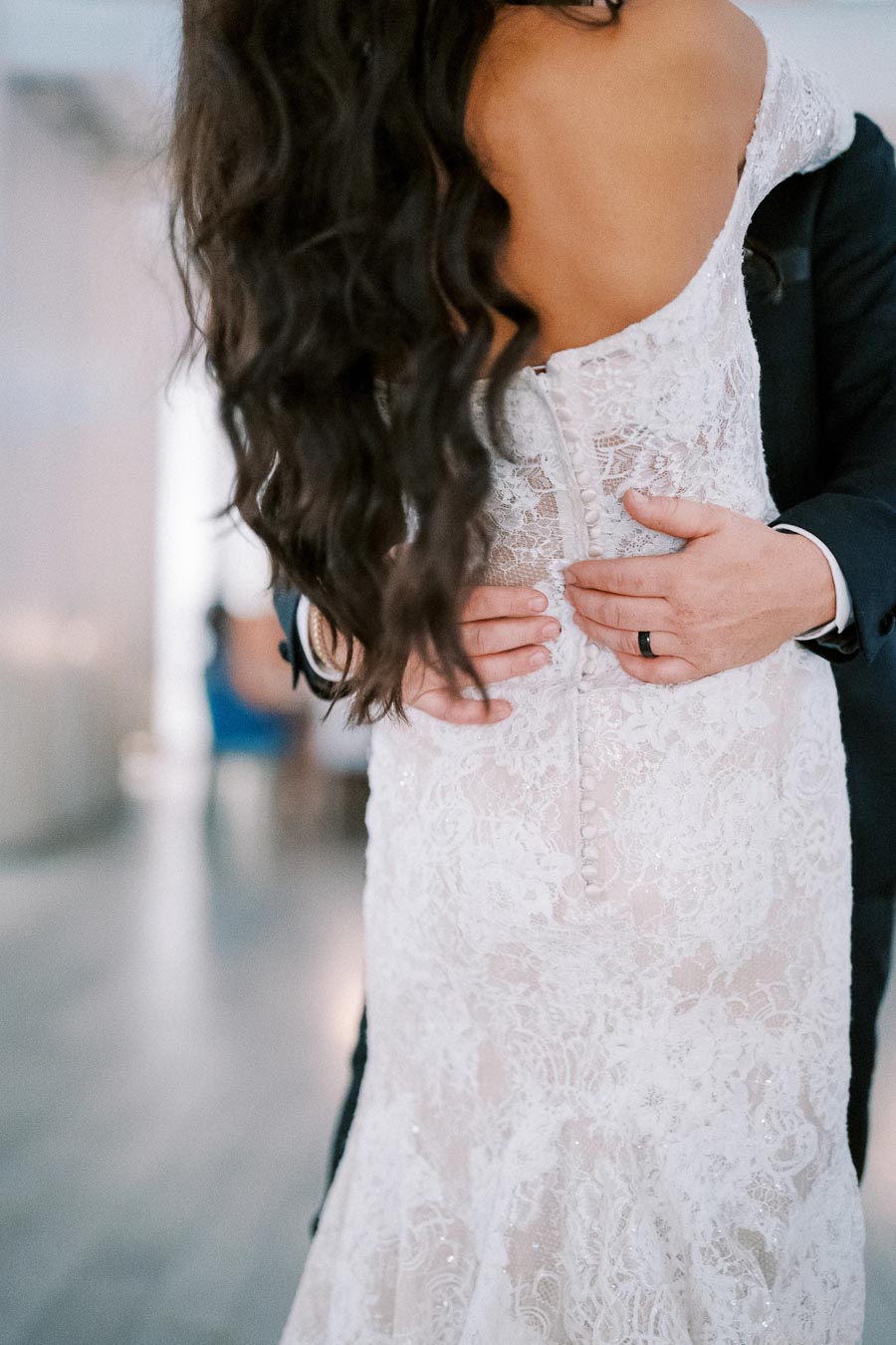 Back view of a bride in a white lace wedding dress being embraced by the groom, showcasing long wavy hair and elegant bridal fashion.