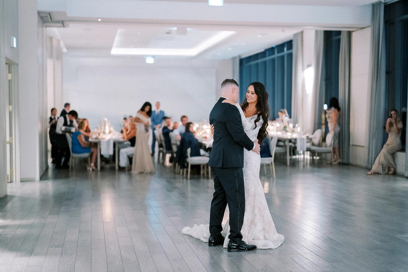 A newlywed couple shares their first dance in a beautifully decorated wedding reception hall, with elegantly dressed guests seated at tables, creating a romantic and joyful atmosphere.