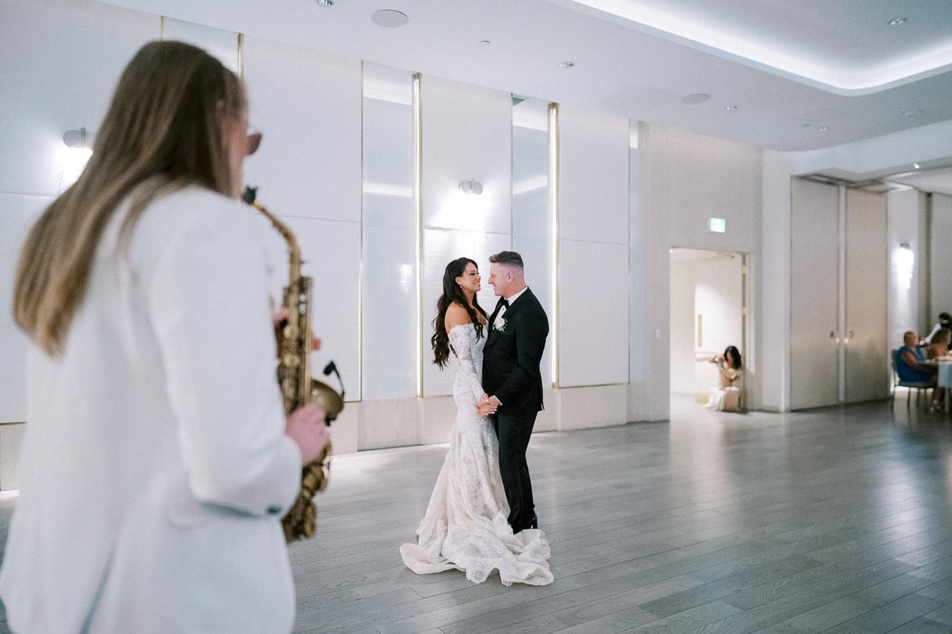 A bride and groom enjoy a romantic first dance in a modern, elegant venue, while a musician plays a saxophone nearby.