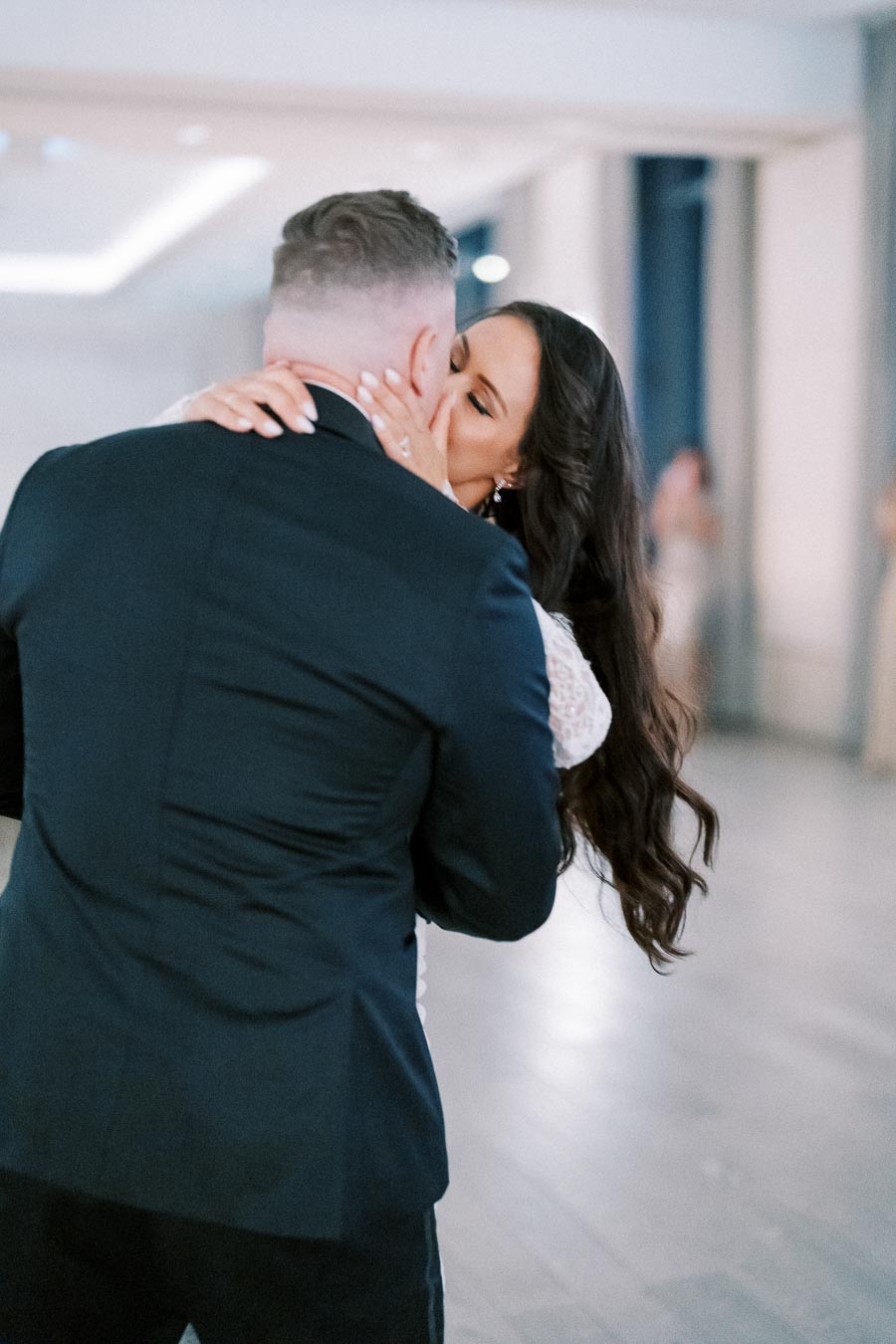 A couple sharing a romantic kiss during their wedding reception, with the bride in a white dress and the groom in a dark suit, captured in a softly lit venue.