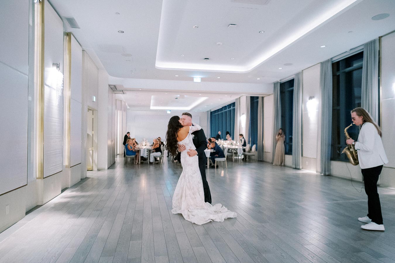 A newlywed couple shares their first dance in an elegant reception hall, accompanied by a live saxophonist, while guests watch seated at tables.